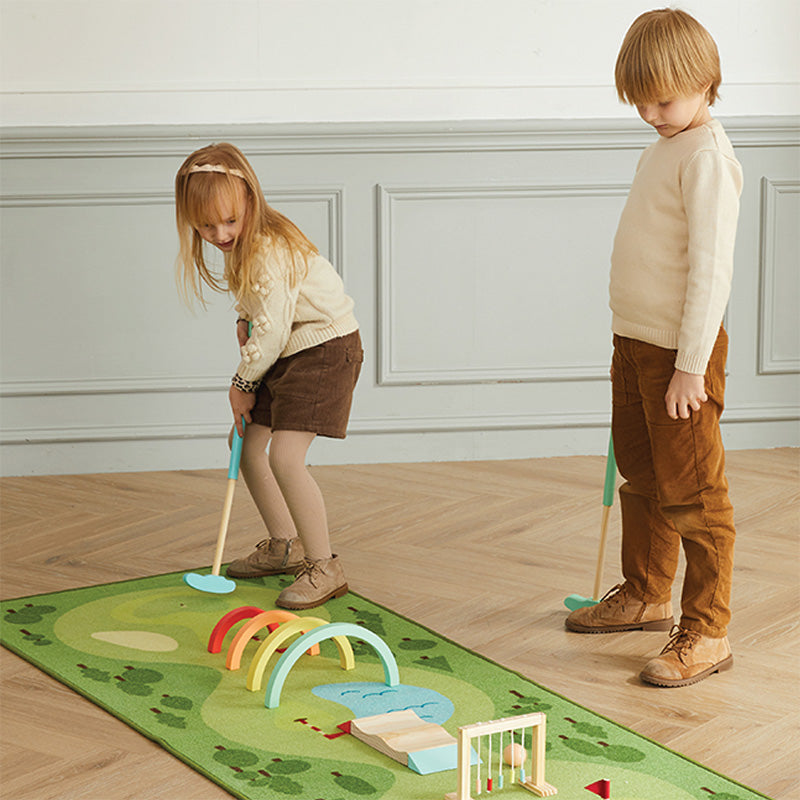 Two children playing with a golf set on a green mat indoors.