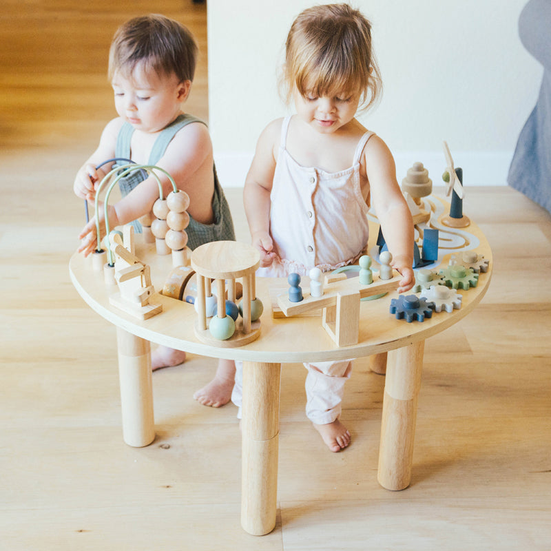 Two toddlers playing with wooden toys at a round natural wood activity table
