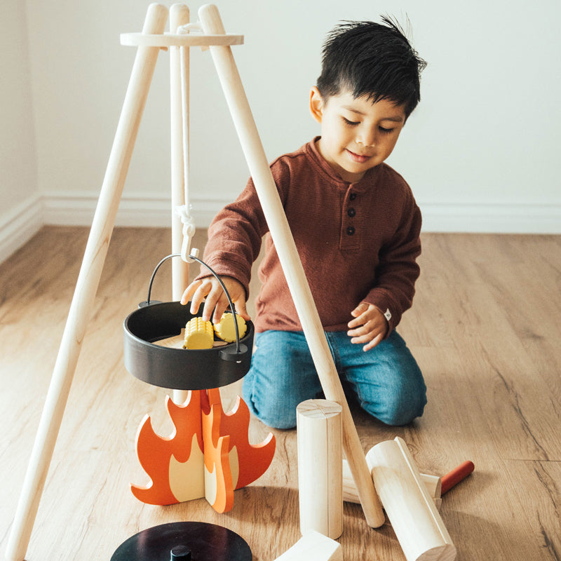 Child playing with toy camping set featuring wooden tripod, black cookware, and flame design