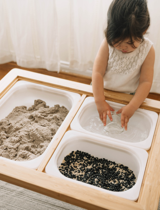 A toddler girl in a white sleeveless top plays with a Montessori-style sensory table featuring sand, water, and black beans in a light wood tray.