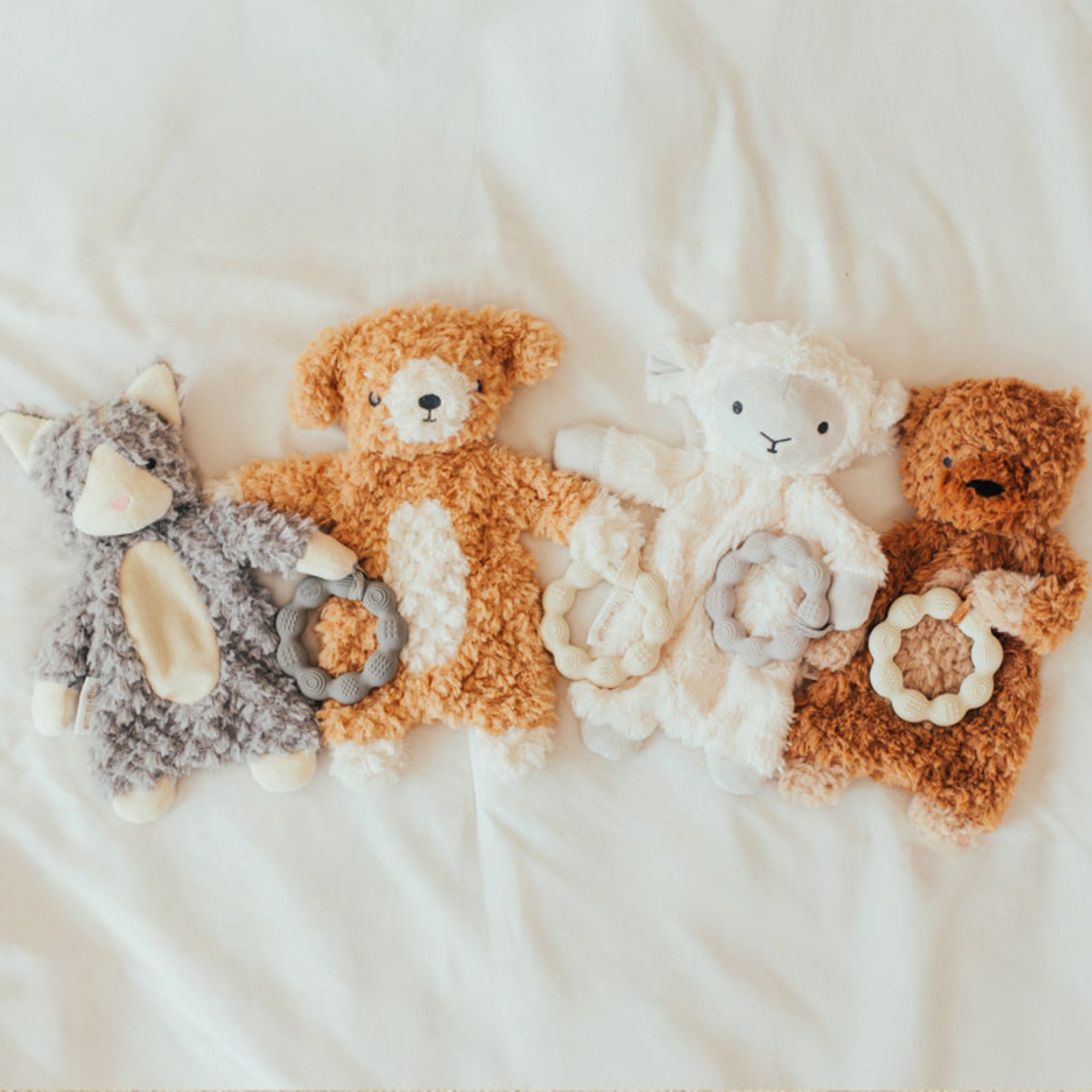 Soft baby lovey stuffed animals in gray, white, and tan colors arranged on white bedding