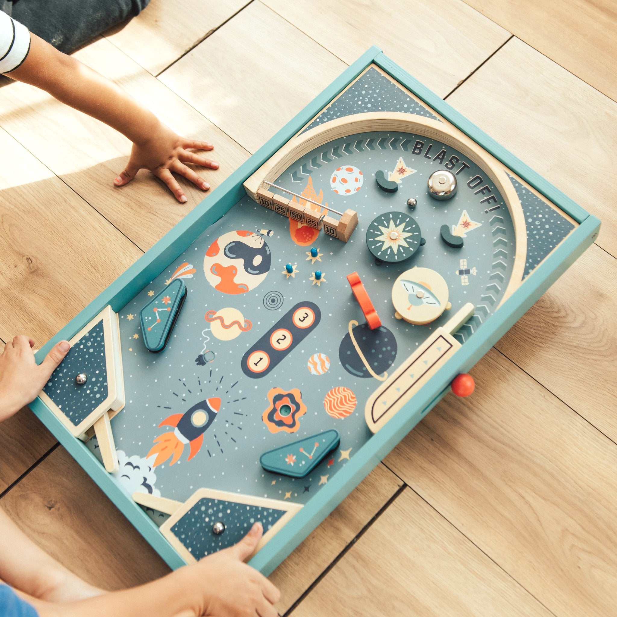 Child's hands on the flippers of the Blast Off Pinball game on a wood floor, seen from above