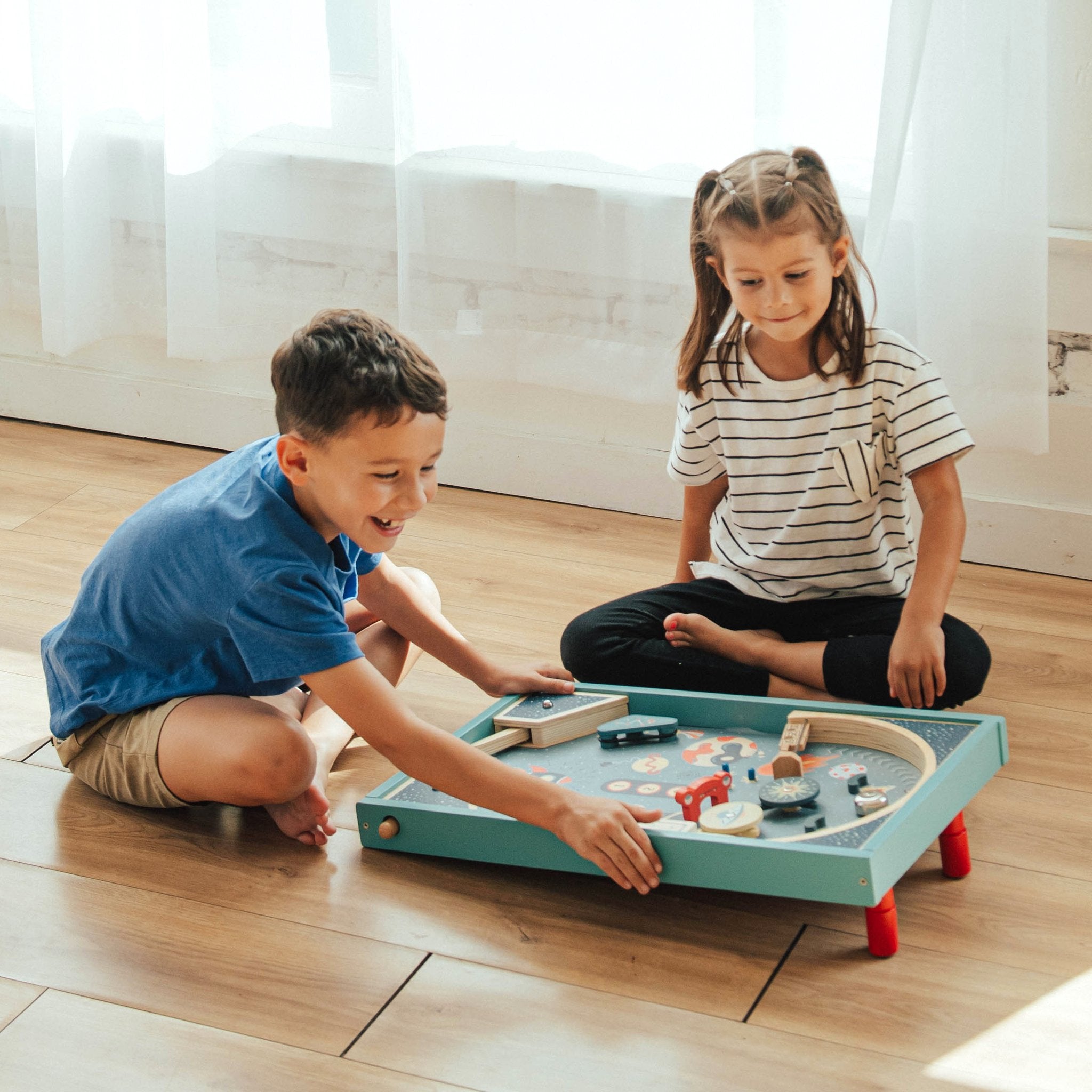 Boy pulling the launcher on Blast Off Pinball while a girl watches, both sitting on a wood floor