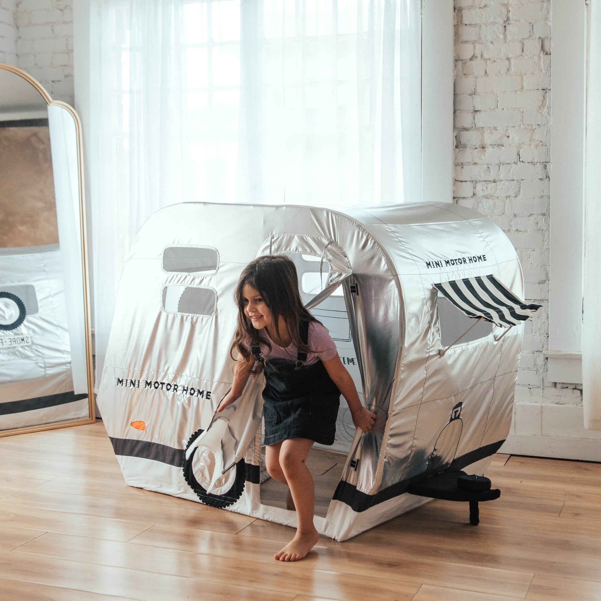 Young girl stepping out the roll-up door of a silver Mini Motor Playhome camper play tent in a sunlit playroom