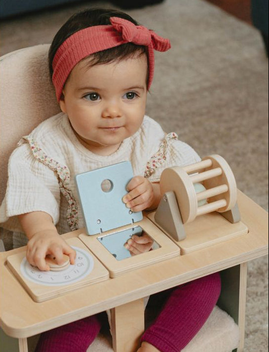 A baby sitting in a high chair using a wooden Montessori activity table with various interactive wooden toys, wearing a pink headband and a white dress.