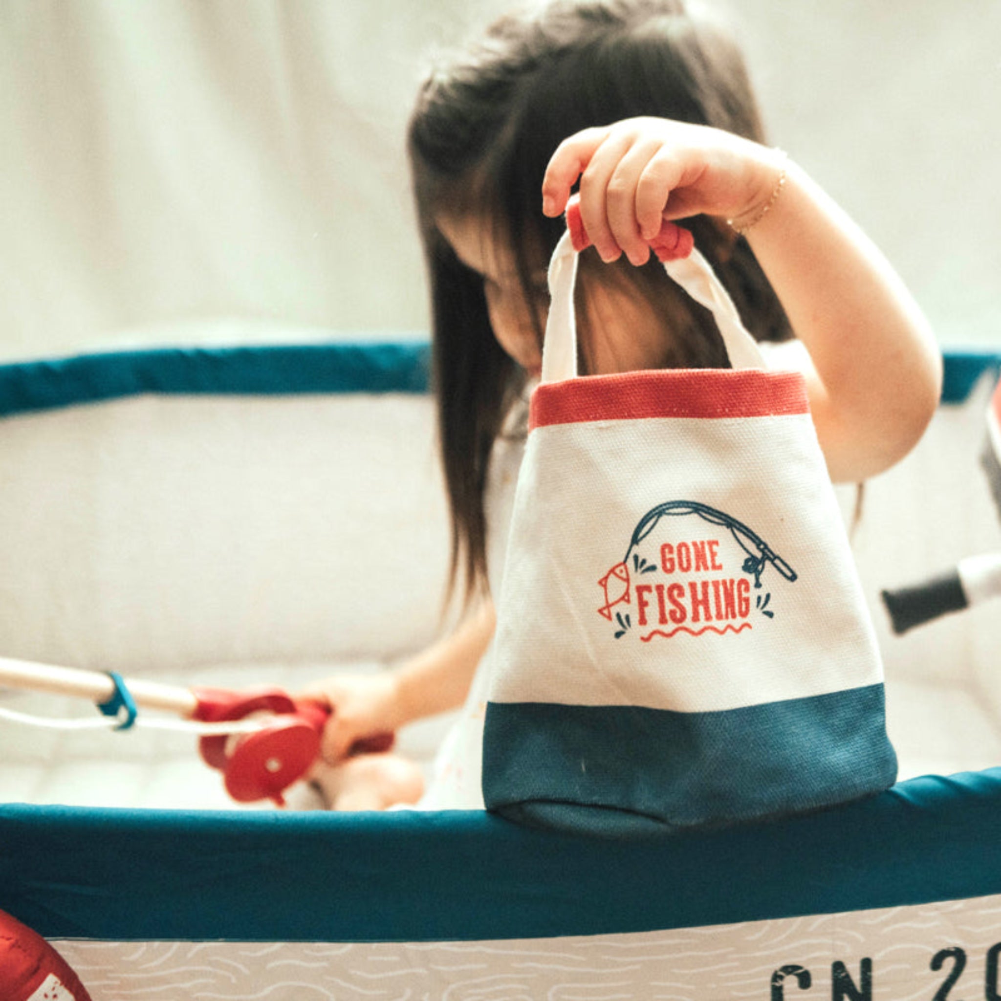 Toddler holding the Gone Fishing canvas bucket by its handle while sitting in a play boat with the wooden fishing rod beside her