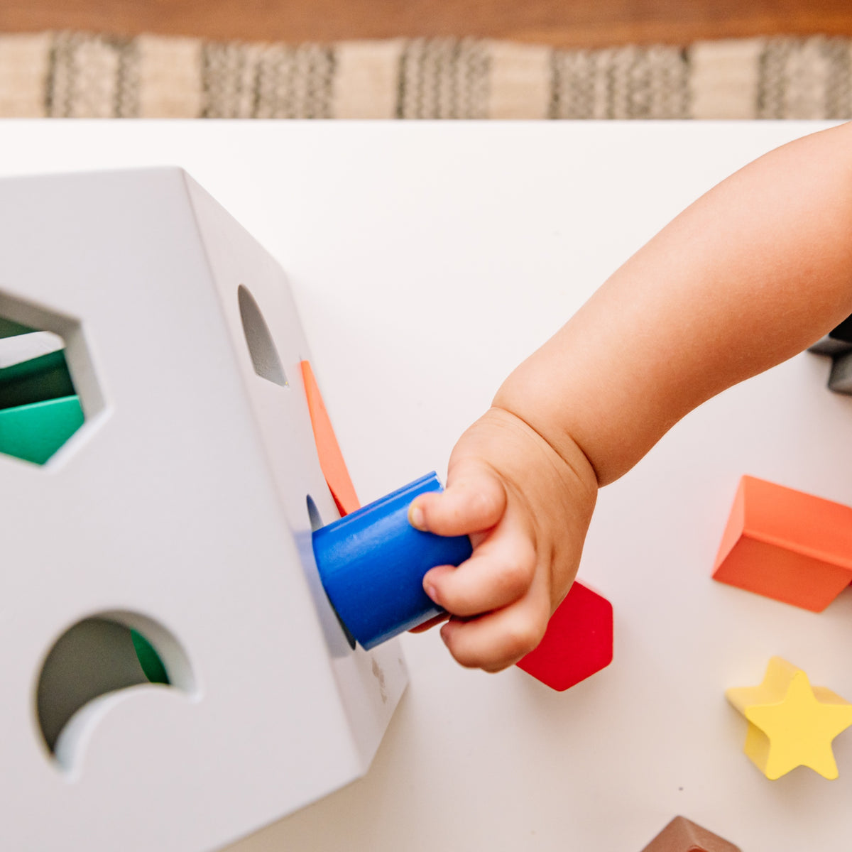Close-up of a toddler’s hand pushing a blue wooden cylinder into the matching opening on the Wonder & Wise Hip Shape Wood Sorter, with red, orange, and yellow blocks nearby.