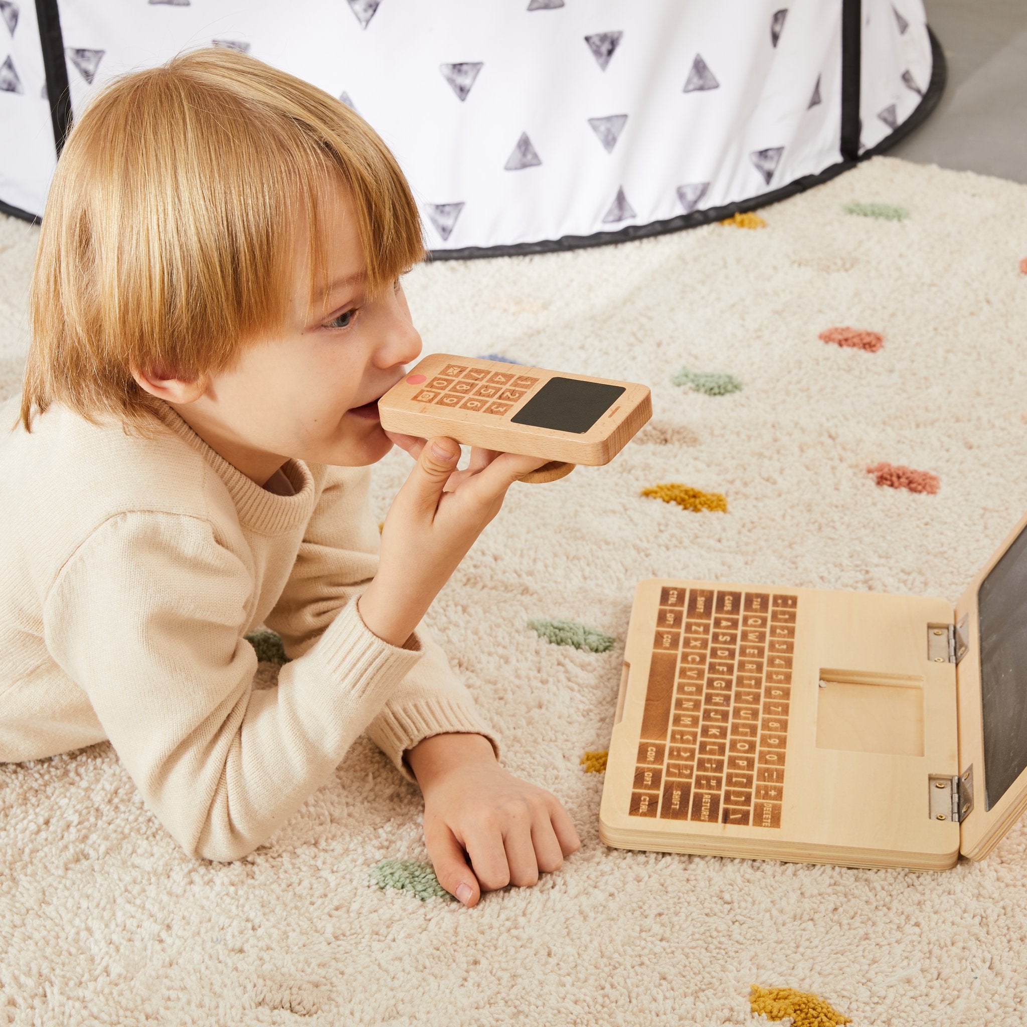 Child lying on a dotted rug holding My Own Cell Phone to their ear, with a wooden play laptop open nearby
