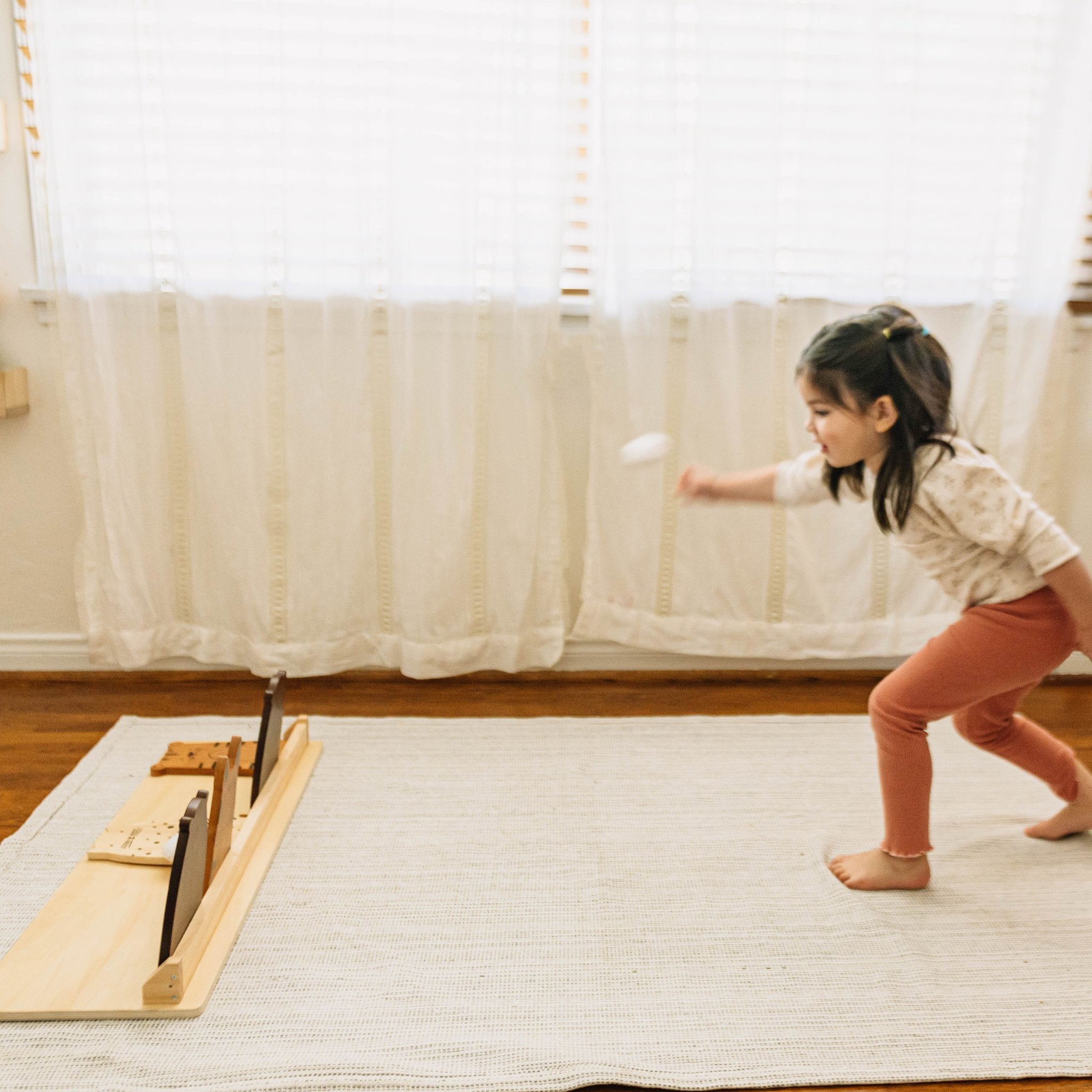 Child in pink leggings tosses a white bean bag at the Zoo Crew Bean Bag Toss on a cream rug by a sunny window